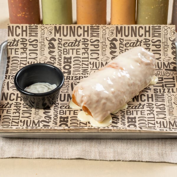 A churro cheesecake, drizzled with white frosting, and a side of cream on a patterned tray.